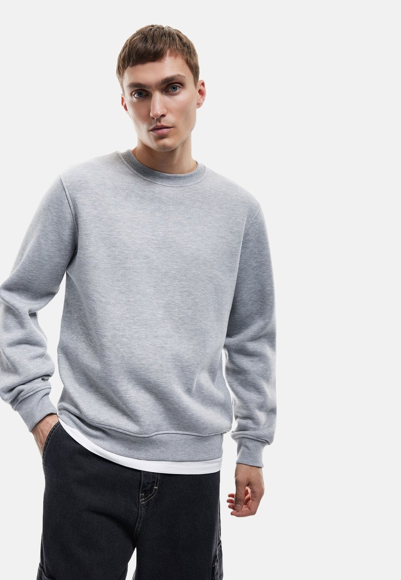 Sudadera gris con cuello redondo y puños acanalados. Presenta una textura suave, mangas largas y un corte relajado, superpuesta sobre una camisa blanca.