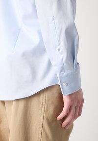 Light blue dress shirt with long sleeves and buttoned cuffs, featuring a clean design and smooth, woven texture. Khaki pants partially visible.