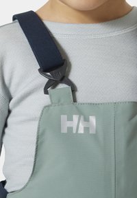 Groene waterdichte overalls met een zilveren logo, verstelbare marineblauwe bandjes en een soepele textuur. Zichtbare schouderbevestigingsclips.