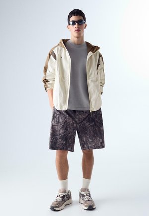 Mladý muž v béžovo-hnedej bunde s kapucňou, šedej tričku, kraťasoch s maskáčovým vzorom, béžových teniskách, bielych ponožkách a čiernych slnečných okuliaroch stojí s rukami v bodoch.