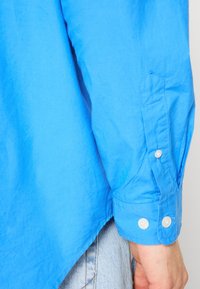 Poignet de chemise à manches longues bleu clair avec deux boutons blancs, portée non rentrée sur un jean en denim clair, avec une main détendue le long du corps.