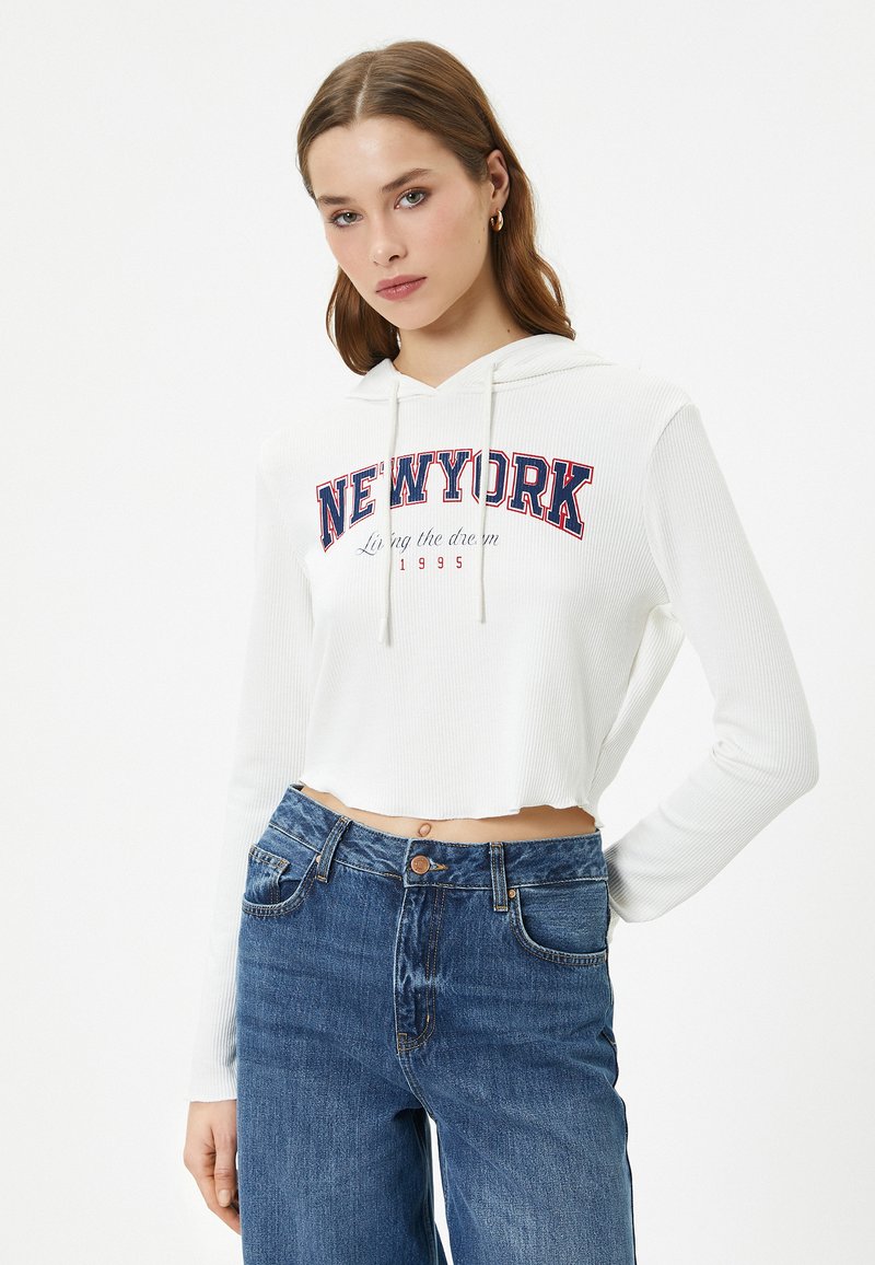 Witte cropped hoodie met ribbelstructuur, voorzien van een "NEW YORK" grafiek in rood en marineblauw. Gecombineerd met blauwe high-waisted jeans.