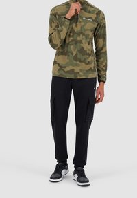 Grüner und beiger Camo-Fleece-Zipper, kombiniert mit schwarzen Cargo-Hosen mit Seitentaschen und schwarzen Sneakers mit grauen Akzenten.