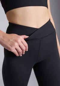 Leggings noirs taille haute avec un design de croisement unique à la taille, en matériau lisse et extensible avec des coutures plates.