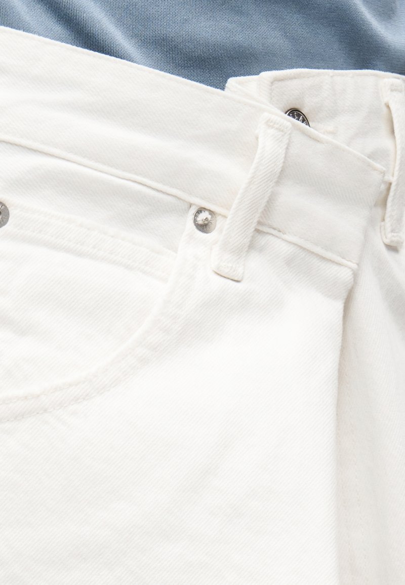 Gros plan sur un jean en denim blanc montrant la ceinture, les passants de ceinture, les rivets en métal, la poche avant et le tissu bleu en dessous.