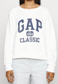 Sweatshirt blanc avec l'inscription "GAP CLASSIC" en bleu marine et le détail "établi en 1969", avec un design court et des poignets côtelés.