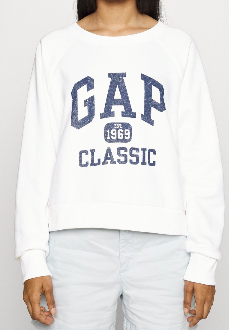 Sweatshirt blanc avec l'inscription "GAP CLASSIC" en bleu marine et le détail "établi en 1969", avec un design court et des poignets côtelés.