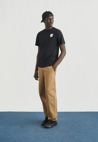 Μαύρο βαμβακερό μπλουζάκι με μικρό λευκό λογότυπο, συνδυασμένο με καμηλό παντελόνι cargo και μαύρα αθλητικά παπούτσια. Το μοντέλο στέκεται σε γαλάζιο χαλί.