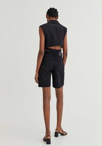 Gilet nero corto con colletto, abbinato a pantaloni corti di jeans neri, con vita alta e orlo arrotondato, posto su uno sfondo neutro.