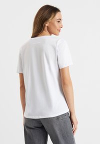 Witte katoenen t-shirt met een ronde halslijn en korte mouwen, voorzien van een klein merkembleem op de achterkant. Model draagt een grijze spijkerbroek.