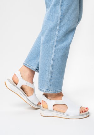 Person wearing light blue jeans and white open-toe sandals with cushioned soles and ankle straps, standing on a white surface.
