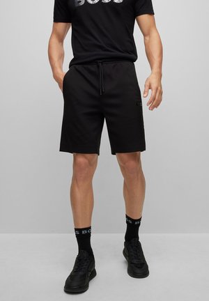 Homme portant un short noir avec cordon, des chaussettes noires et des baskets noires, debout devant un fond gris uni.