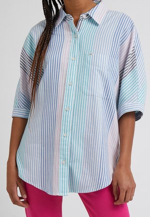 Kobieta nosząca luźną koszulę na guziki w pasy w pastelowych odcieniach niebieskiego, fioletowego i turkusowego z rękawami do łokcia oraz różowe spodnie.