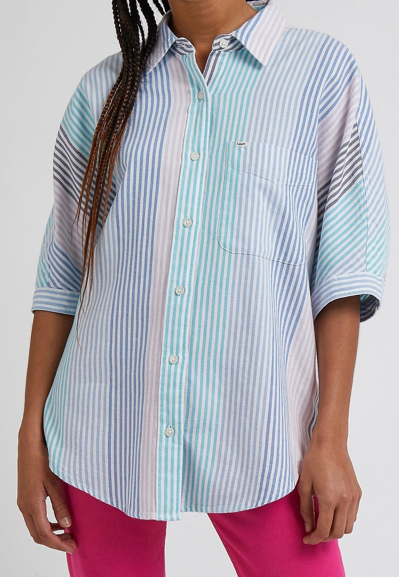 Femme portant une chemise ample à rayures pastel bleu, violet et turquoise avec des manches trois-quarts et un pantalon rose.