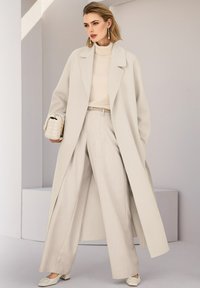 Lange, licht beige jas over een bijpassende coltrui en ruime broek, met een gestructureerde handtas en schoenen met blokhak.