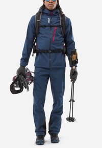 Veste d'extérieur navy avec des accents rouges, pantalons navy assortis, portant des raquettes à neige et des bâtons de randonnée, vêtue de gants et d'un sac à dos.