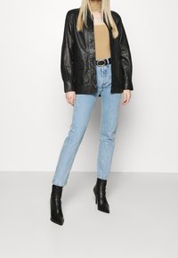 Veste en cuir noire à boutons, jean bleu clair et bottines noires à talons. La tenue comporte un haut beige en dessous.