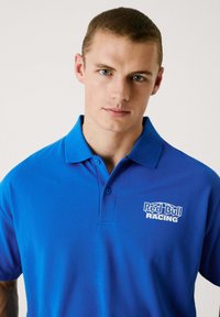 Blå pikétröja i texturerad tyg, med krage, två knappar och en vit "Red Bull Racing"-logotyp på vänster bröst.