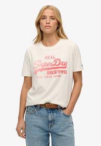 Biała bawełniana koszulka z różowym nadrukiem "SuperDry", okrągły dekolt, krótki rękaw, w połączeniu z niebieskimi dżinsami i plecionym brązowym paskiem.