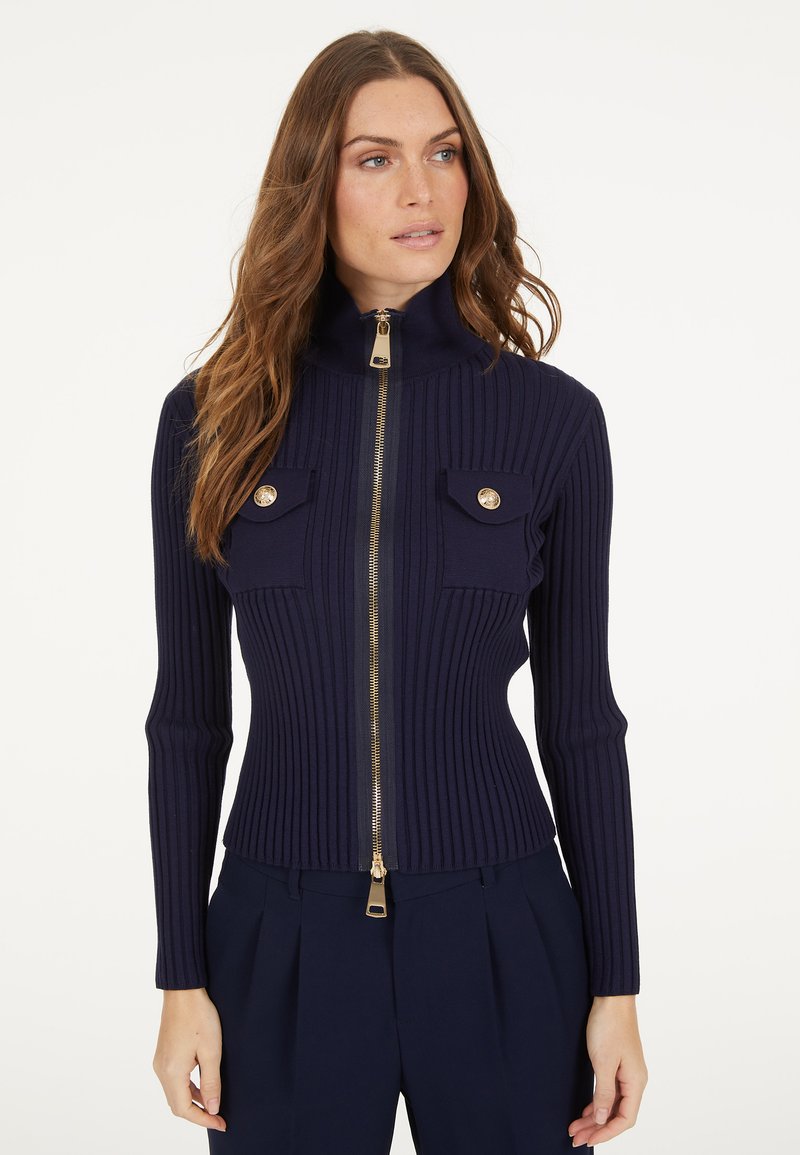 Marineblaues geripptes Strickjacke mit hohem Kragen, goldfarbenem Reißverschluss und Fronttaschen, die mit goldenen Knöpfen verziert sind. Taillierte Passform, lange Ärmel.