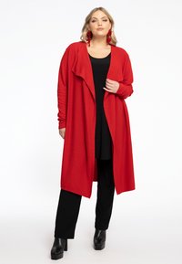 Cardigan long rouge avec un devant drapé et ouvert, fabriqué dans un tissu doux, associé à un haut noir et un pantalon, et des bottes à plateforme courtes noires.