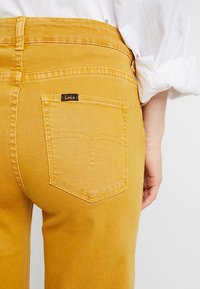 Pantalones de mezclilla amarillos con cintura alta, bolsillo trasero con una etiqueta negra "Lois" y costuras en contraste. Textura suave y diseño ajustado.