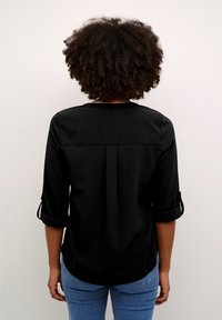 Blouse noire à manches trois quarts et poignets retroussés. Texture lisse, design simple et détail plissé dans le dos.
