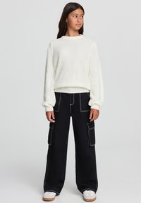 Biały sweter dzianinowy o fakturze ściągacza, zestawiony z czarnymi spodniami cargo o szerokich nogawkach z kontrastowymi szwami i bocznymi kieszeniami. Białe trampki.