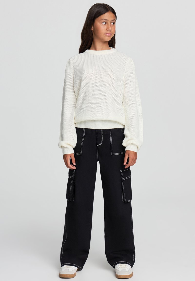 Biały sweter dzianinowy o fakturze ściągacza, zestawiony z czarnymi spodniami cargo o szerokich nogawkach z kontrastowymi szwami i bocznymi kieszeniami. Białe trampki.