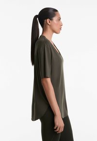 Olivengrøn kortærmet bluse med V-hals, lavet af glat stof, har en afslappet pasform og en længere ryg. Båret over sorte leggings.
