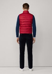 Chaleco acolchado rojo con costuras horizontales, cuello alto y textura suave, llevado sobre una camisa de manga larga azul marina y pantalones negros.