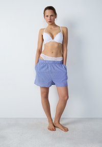 Biały stanik z koronkowym wykończeniem noszony z niebieskimi spodniami w paseczki. Spodnie posiadają elastyczny pas i boczne kieszenie. Gładka tekstura materiału, swobodny krój.