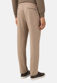 Svetle béžové kalhoty s hladkou strukturou, elastickým pasem a jednou zipovou kapsou na zadní straně. Stylizováno s tmavými teniskami.