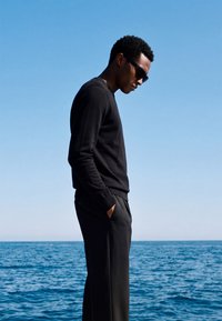 Homme vêtu de vêtements noirs et de lunettes de soleil, debout les mains dans les poches, regardant vers le bas près d’une mer calme sous un ciel bleu clair.