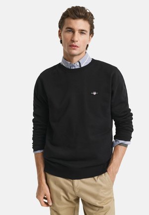 Felpa nera con collo a giro, polsini e vita a costine. Presenta un piccolo logo stampato sul petto. Indossata sopra una camicia button-up azzurra chiara.