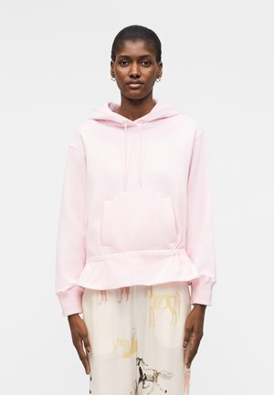 Sieviete valkā gaiši rozā kapuciņu ar priekšējo kabatu un krēmkrāsas plašas bikses ar zirgu apdruku.