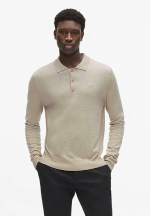 Mees, kes kannab beežikat pika varrukaga polosärki ja musti pükse, seisab käed taskutes ühevärvilise valge tausta ees.