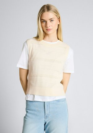 Donna bionda che indossa un maglione senza maniche lavorato a maglia color crema sopra una t-shirt bianca e jeans azzurri, in piedi davanti a uno sfondo grigio uniforme.