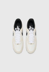 Nike Air Force 1 sneakers in cream and white leather with black accents, featuring perforated toe boxes and white laces. Black sock lining included.
