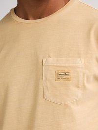 Tricou bej cu mânecă scurtă, din bumbac, cu un buzunar pe pieptul stâng, având o etichetă de marcă. Textură moale și netedă.