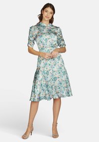 Frau in einem knielangen Blumenkleid mit gerafften Ärmeln, die mit sanft gefalteten Händen dasteht und leichte Absatzsandalen trägt.