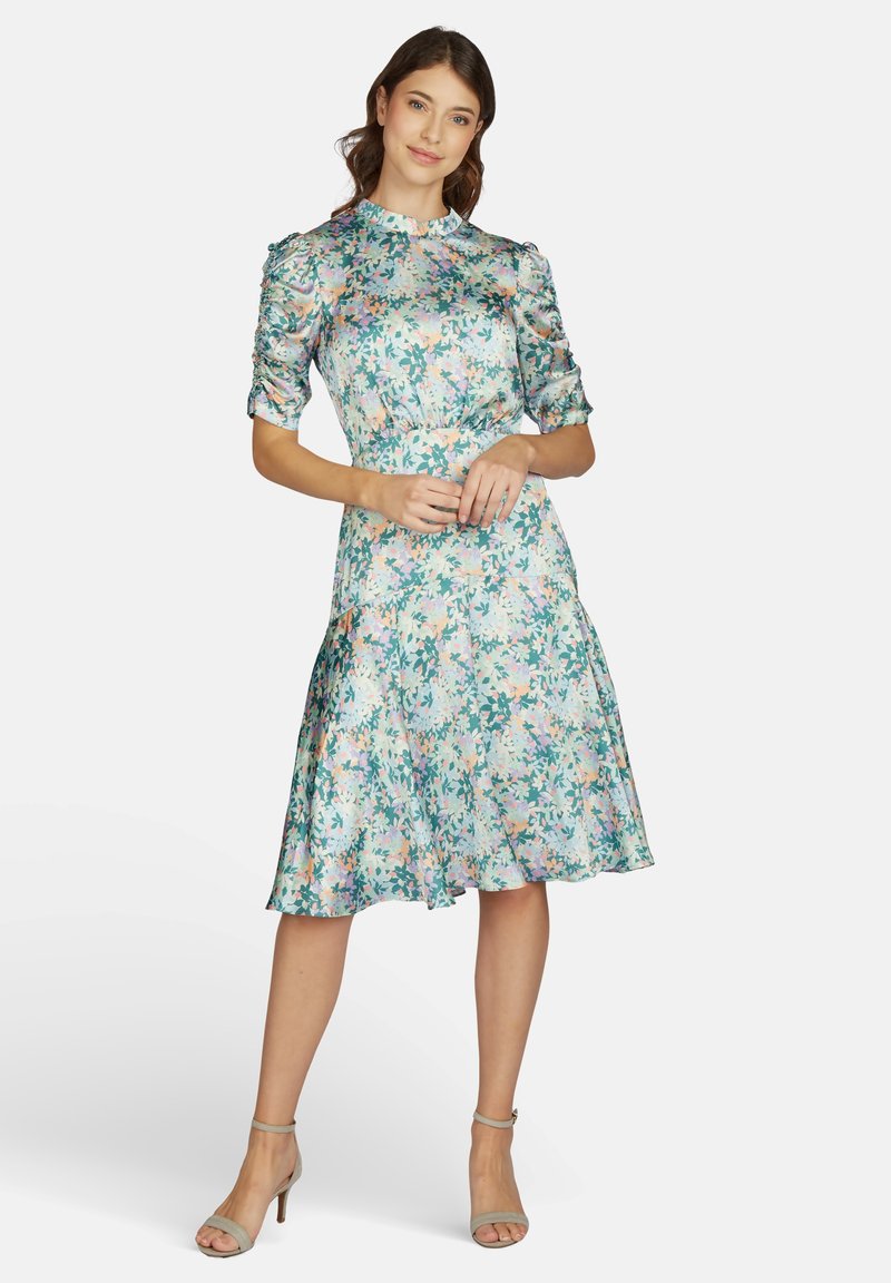 Frau in einem knielangen Blumenkleid mit gerafften Ärmeln, die mit sanft gefalteten Händen dasteht und leichte Absatzsandalen trägt.