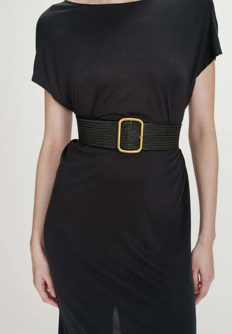 Robe noire à manches courtes avec une large ceinture noire ornée d'une grande boucle carrée dorée à la taille, portée par une personne debout.