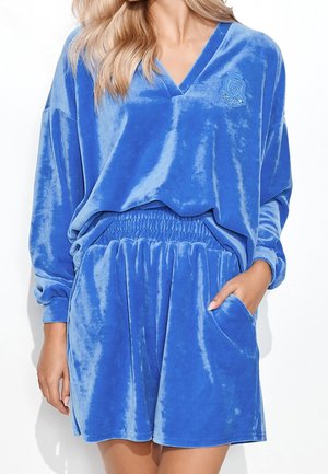 Kobieta ubrana w luźną, niebieską welurową bluzkę z długim rękawem i dekoltem w kształcie litery V oraz pasujące krótkie spodenki z elastycznym stanem i bocznymi kieszeniami.