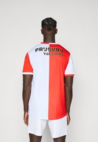 Rotes und weißes Sporttrikot mit kurzen Ärmeln und abgerundetem Saum. Text "PRUSVR'J vakanties" in Schwarz, positioniert auf dem oberen Rücken.