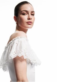 Vrouw met slicked-back haar, draagt gouden ringoorbellen en een witte off-shoulder top met oogjes kantdetails, kijkt naar de camera.