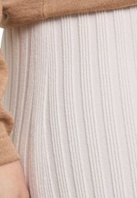 Beżowy sweter z dzianiny na tle fakturowanej, żeberkowanej jasnoróżowej tkaniny. Materiał wygląda na miękki, z delikatnymi pionowymi liniami i połyskiem.