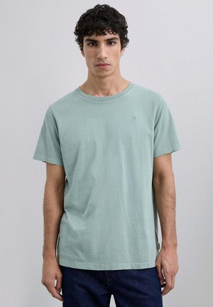 Camiseta de algodón verde menta clara con cuello redondo y mangas cortas, que presenta un pequeño detalle de logo en el pecho. Diseño simple y casual.