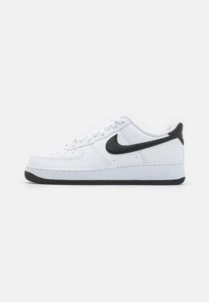Bílé kožené tenisky s černým logem Nike Swoosh, kulatá špička, šněrovací přední část, strukturovaná černá gumová podrážka a reliéfní nápis "Air" na patě.