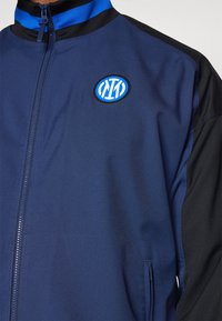 Navy blue zip-up jacket with black accents and a blue circular logo. Features ribbed collar and smooth fabric texture, with zippered pocket.